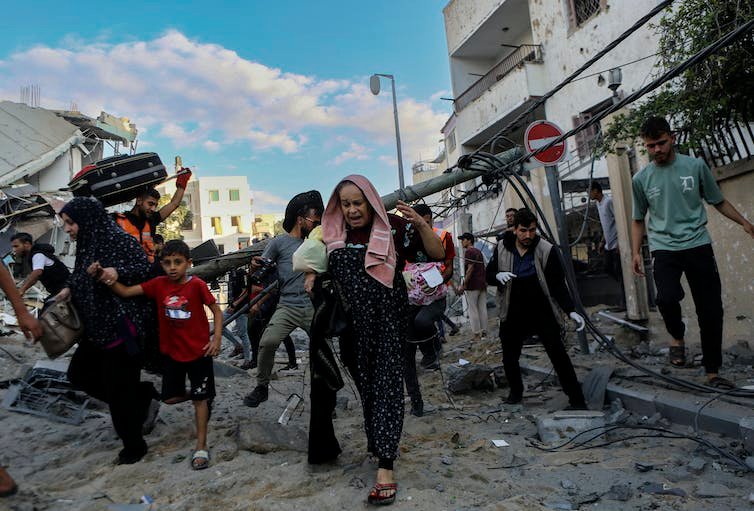 Several people, including women and children, running out of their homes. Behind them are some partially damaged buildings.