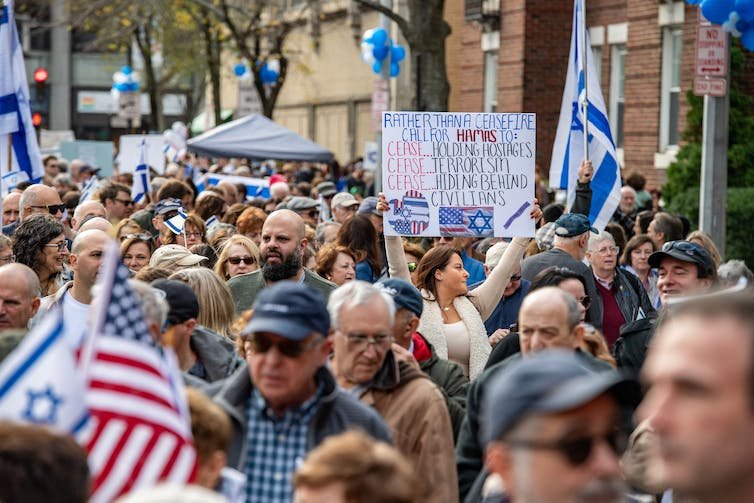 Sekelompok orang berjalan di jalan bersama-sama, memegang bendera Israel dan Amerika Serikat dan sebuah tanda yang bertuliskan, 'Daripada gencatan senjata, serukan agar Hamas berhenti menyandera, hentikan terorisme, hentikan bersembunyi di balik warga sipil.'